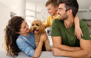 Family of three with a fluffy dog, smiling warmly in a bright living room. The mother holds the dog, while the father and son lean in joyfully.